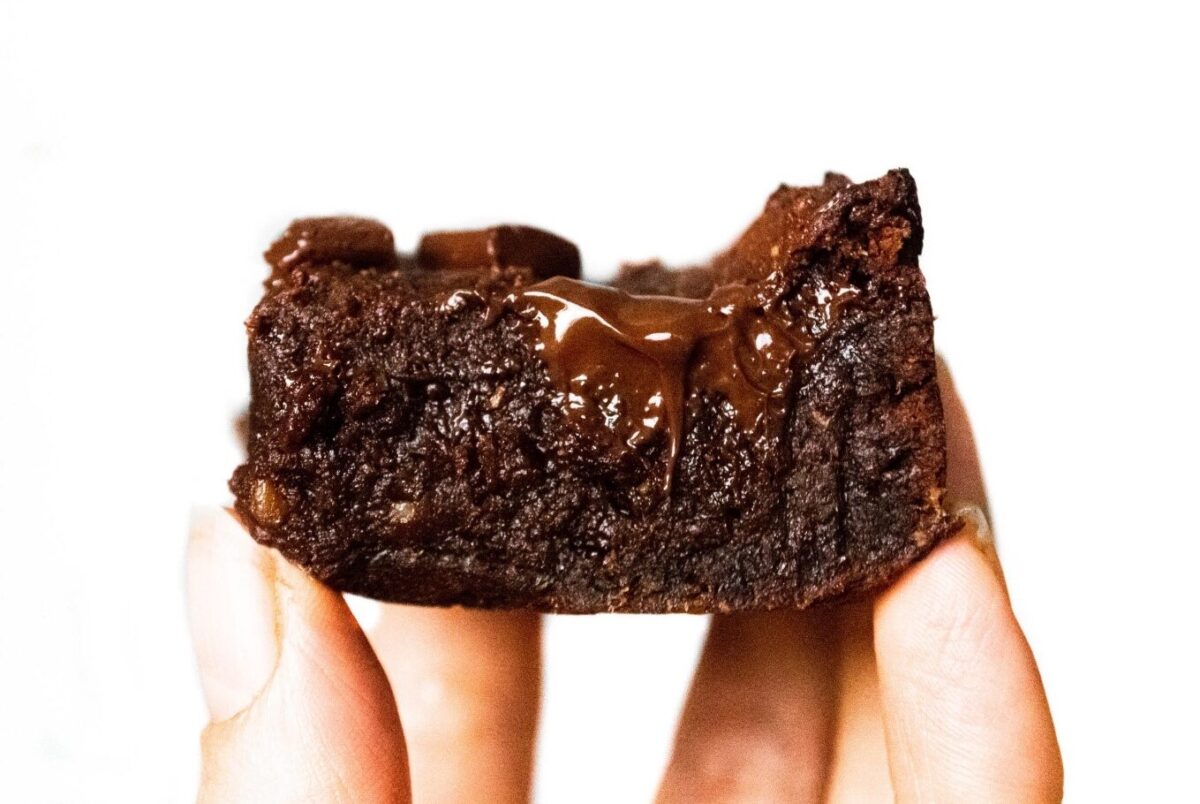 Someone holding up a slice of vegan chocolate brownie in front of the camera, with melted chocolate oozing from the middle.