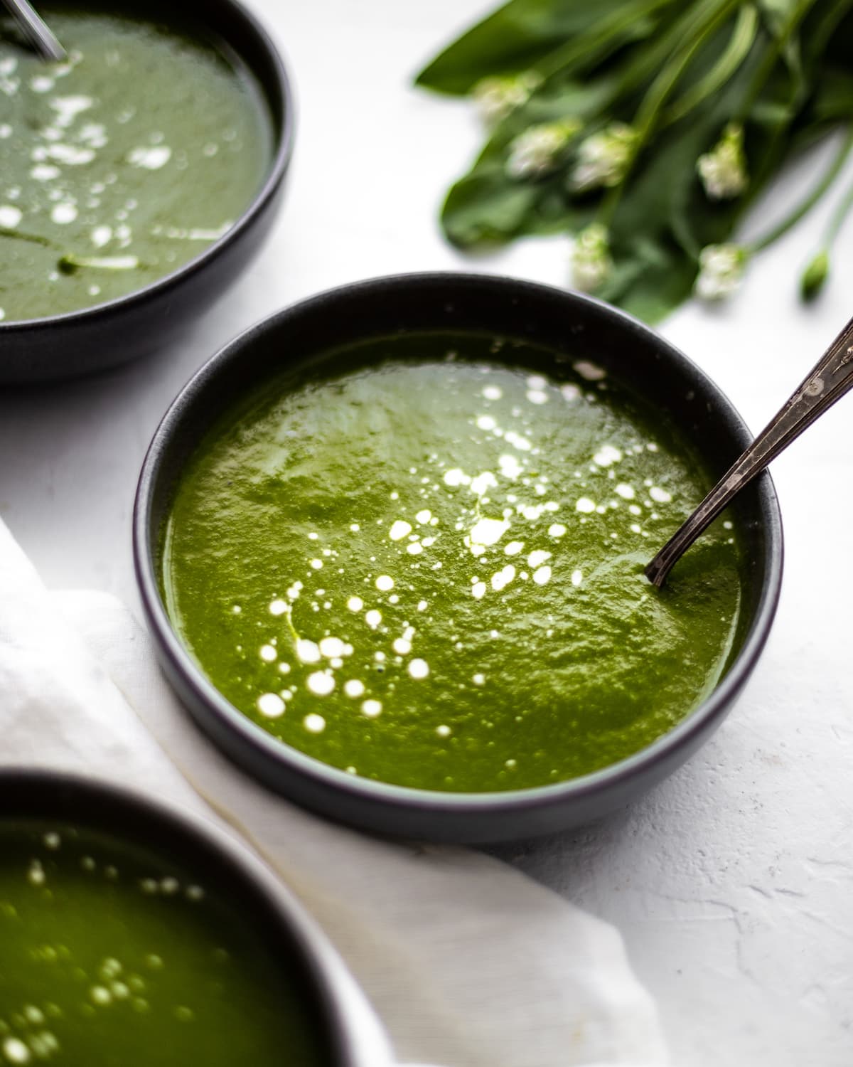 Photo of three bowls of wild garlic soup on a table with a napkin and a bunch of wild garlic. The bowls of soup are splattered across the surface with cashew cream.