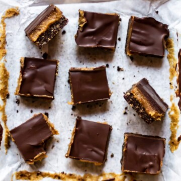 9 vegan caramel slices cut in to squares, on a white sheet of greaseproof paper. Two slices have been turned on their side to show the layers within - the base, caramel filling, and chocolate topping.