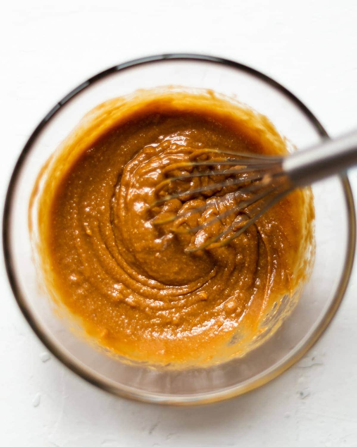 A glass bowl full of Thai style vegan satay sauce, on a white background, with a whisk in the cenre of the bowl.