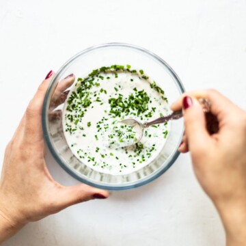 A glass bowl, on a white background, filled with the chive dip being stirred to mix in the freshly chopped chives.