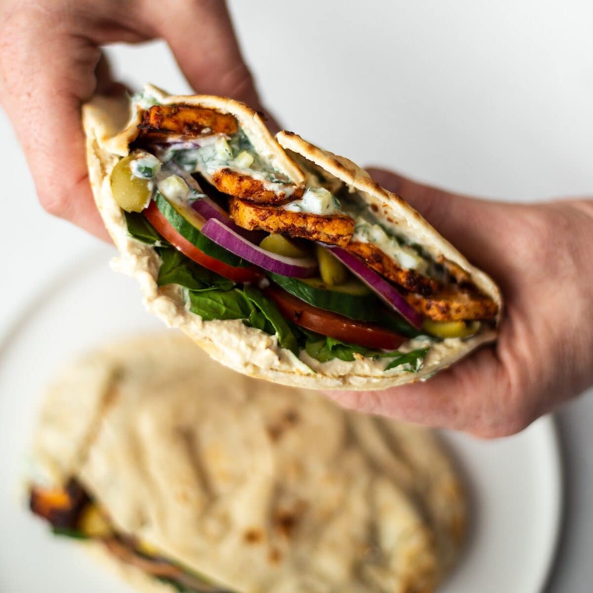 Two hands holding a pitta, loaded with marinated tofu, salad, hummus and tzatziki. Photographed being held over a plate with another filled pitta, in the background, on a plate, out of focus.