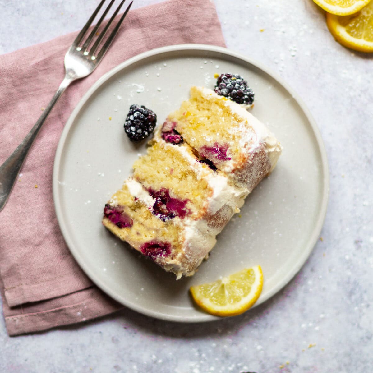 An over head shot of a slice of lemon blackberry cake on a plate. The plate is sat on a pink napkin, with a fork sat next to it.