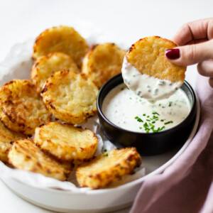 Side angle of a white bowl filled with the baked hash browns, and a black bowl filled with chive dip. One has brown is being dipped ⅓ of the way in to the chive dip.