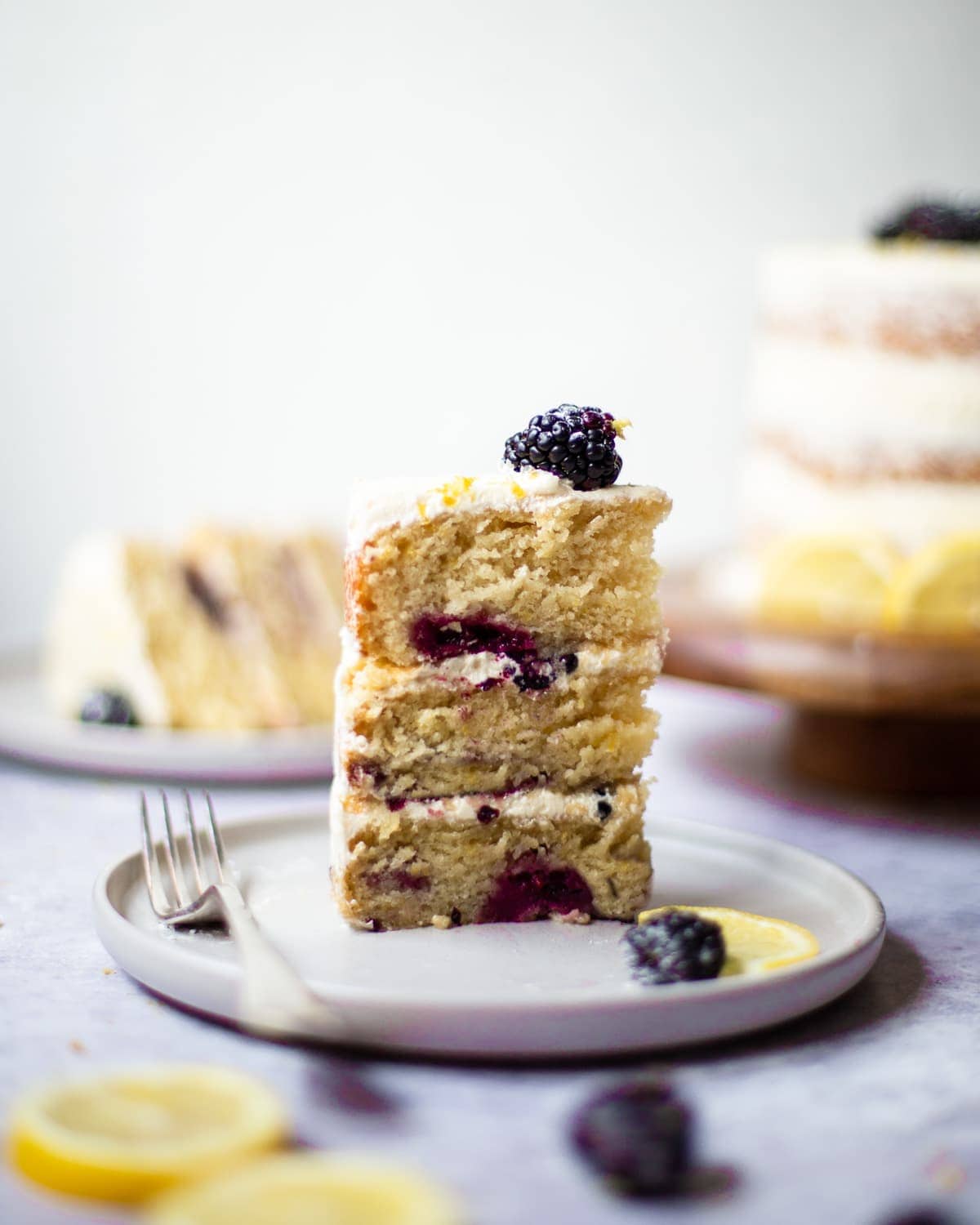 A slice of lemon blackberry cake, stood on a plate with a slice of lemon, with more cake in the background, out of focus.