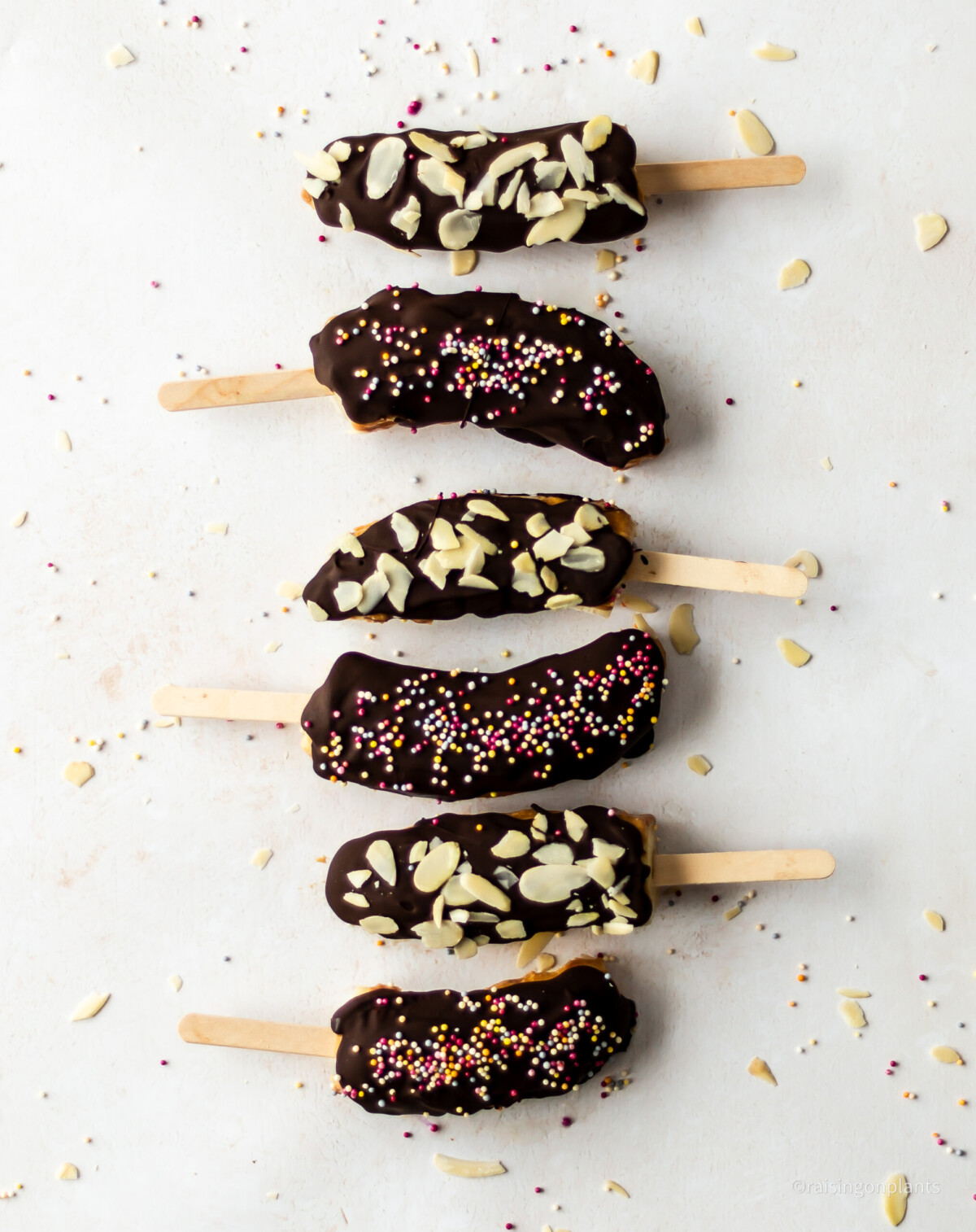 Six chocolate coated banana lollies laid on a white background, with sprinkles and flaked almonds. 