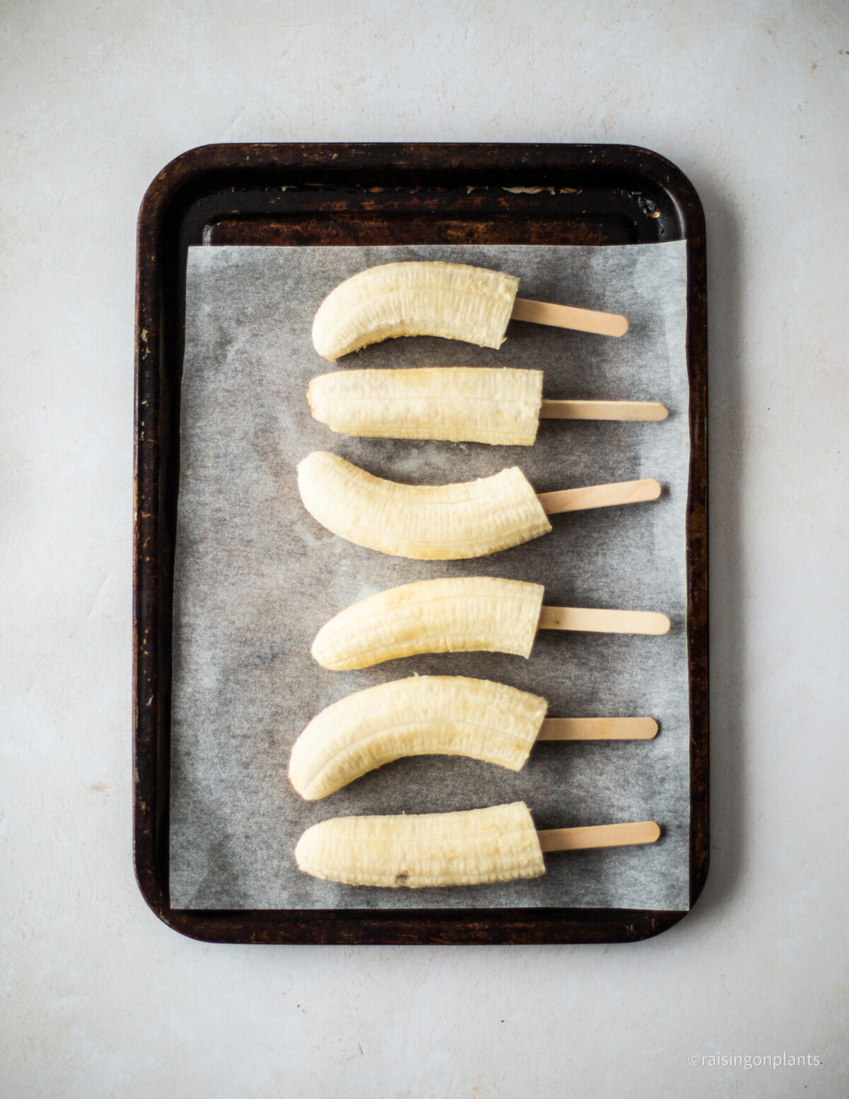 Six banana halves, lined up on a lined baking tray, with ice lolly sticks inserted at one end.